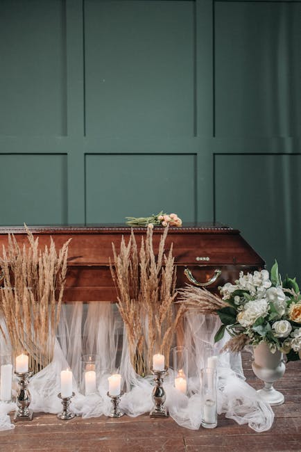 A floral arrangement featuring white roses, peonies, and greenery placed in a white ceramic vase, positioned on a wooden surface with a large bouquet of mixed white flowers and greenery to the right.
