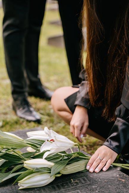 A woman dressed in black is kneeling beside a grave, arranging a floral tribute consisting of white lilies with large, smooth petals and prominent stamens, accented by green leaves and long stems. The