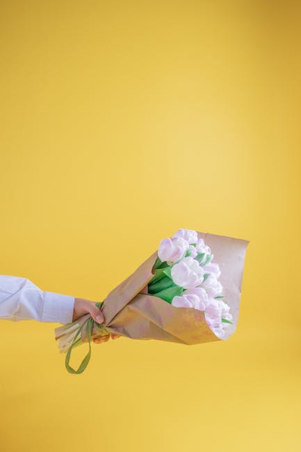 A hand, dressed in a white shirt, holds a bouquet of pale pink tulips wrapped in light brown craft paper with a green ribbon, presented against a vibrant yellow background. The tulips are fresh with s