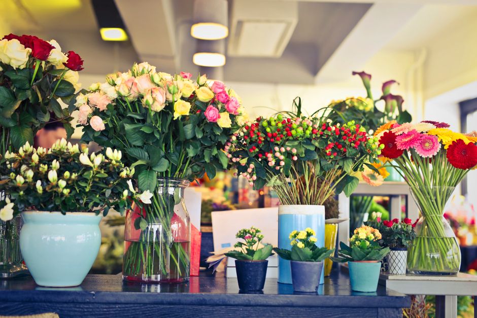 A variety of fresh flower arrangements on display at a floral shop, including pink and yellow roses in clear and blue vases, colorful gerbera daisies, and small potted green plants, set on a dark wood