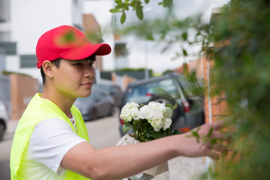 A young male flower delivery courier wearing a red cap and a bright yellow safety vest is placing a bouquet of fresh white hydrangeas against a leafy green wall outside a residential area. The bouquet