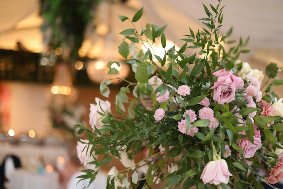 A large floral arrangement featuring soft pink roses with layered petals, complemented by clusters of small pink chrysanthemums and white flowers, all intertwined with lush green foliage and glossy le