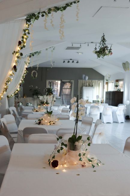 An elegantly decorated indoor event space featuring floral arrangements with white orchids and dark red roses placed on tables with white tablecloths. The room has a soft, ambient glow from string lig