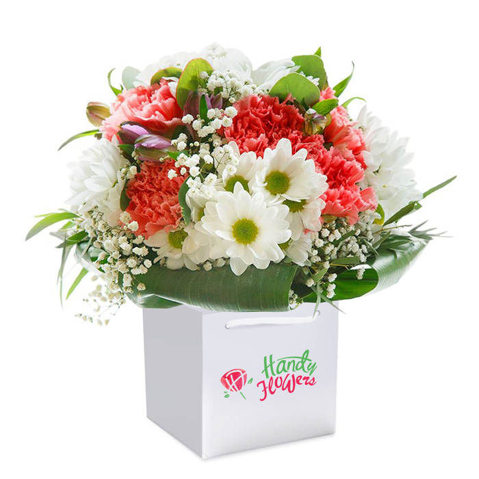 Bright and uplifting, this handcrafted floral arrangement greets the viewer from within a minimalist, white gift bag-practical for easy delivery throughout Woodford's charming streets. At its heart, soft coral-pink carnations display layers of ruffled, velvety petals, their hues gently drifting between pale peach and lively coral, offering a subtle sweet fragrance. Nestled closely beside them, pristine white daisies with bold, lime-green centers add a playful lightness, their petals crisp and joyful like a walk down Snakes Lane in spring. Accents of airy, cloudlike baby's breath (gypsophila) are artfully scattered throughout the bouquet, providing a delicate contrast and a whisper of innocence. A sprinkling of nearly-open purple buds, peeking out from one side, add intrigue and a sense of promise to the gathering of blooms. Encircling all is an abundance of broad, deep green foliage, high-gloss and sturdy, carefully shaped to form a polished collar around the rounded bouquet. The entire arrangement is photographed under gentle, natural light against a seamless white background, underscoring the flowers' freshness and the florist's attention to detail. Perfect for celebrating a loved one or marking a milestone with a taste of Woodford's best floristry, this bouquet is a radiant expression of care, quality, and the joy of giving flowers in a vibrant East London community.